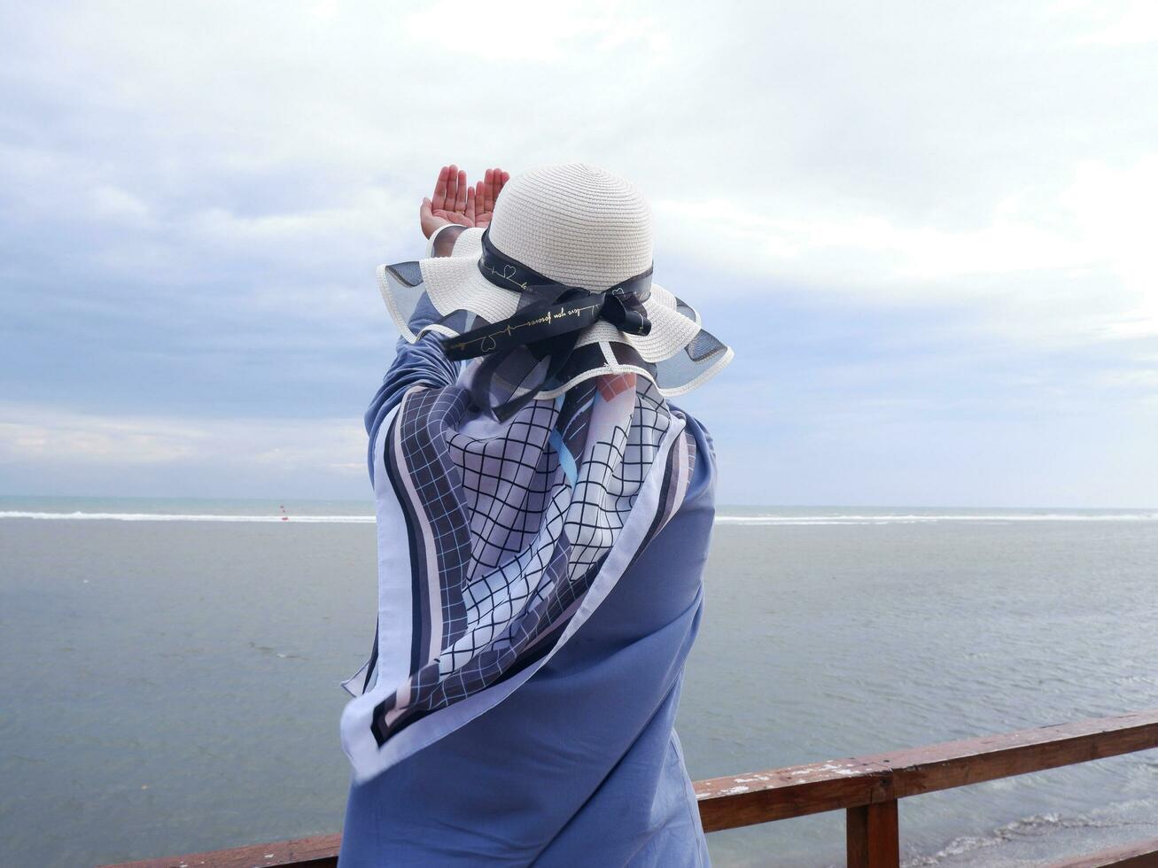 tillbaka av de kvinna i de hatt på de tropisk strand vem är ser på de himmel och de hav medan höjning henne hand i bön foto