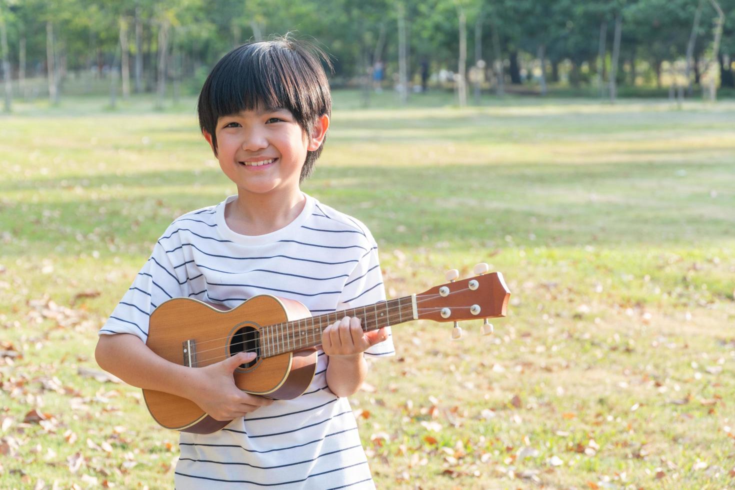 Lycklig asiatisk pojke spelar på ukulele gitarr i de parkera foto
