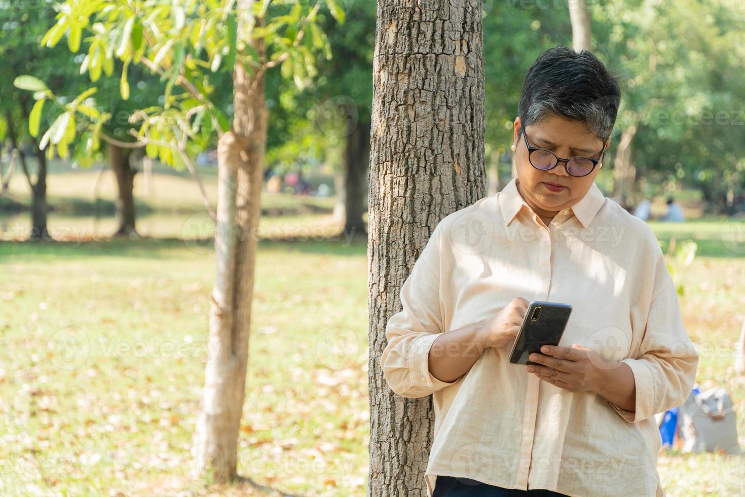 asiatisk senior kvinna stående på träd i de parkera spelar på mobil telefon foto