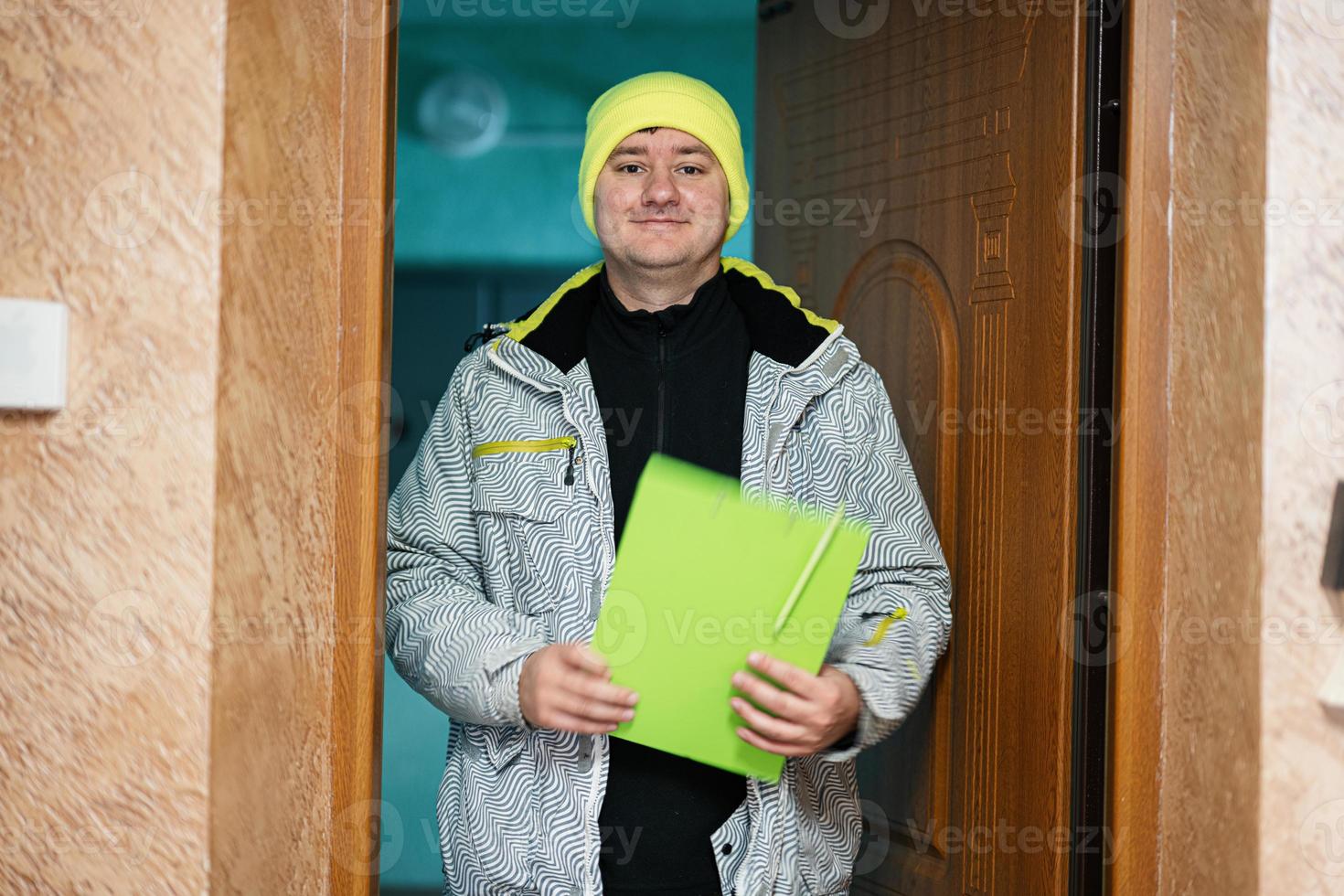 leverans man. kurir i grön Färg hatt innehav en Urklipp medan stående mot dörr av bostads- hus foto