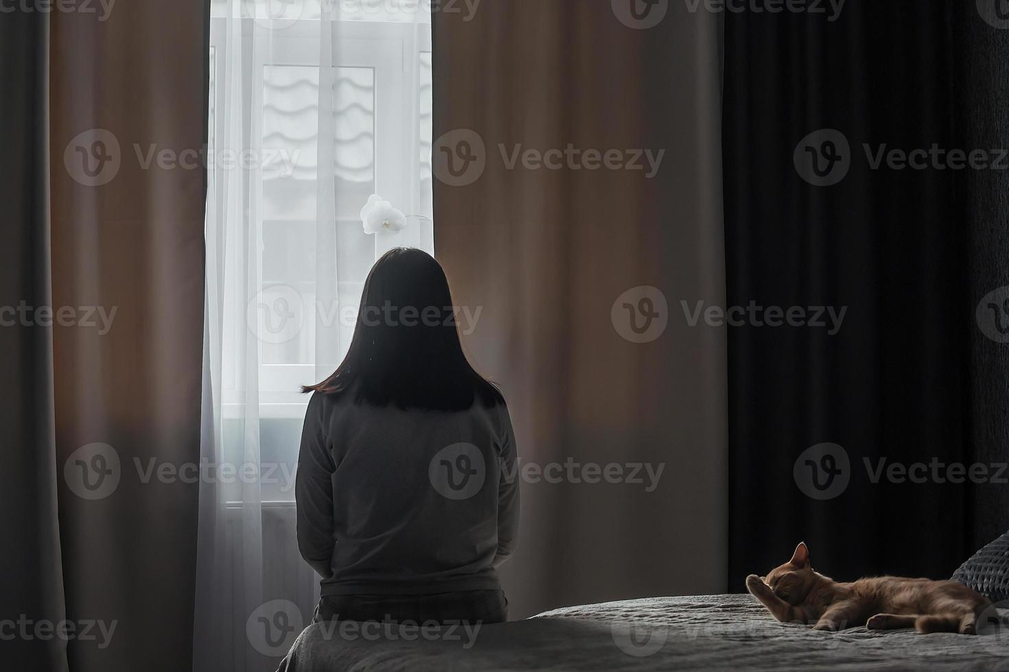 silhuett av en ung kvinna i pyjamas mot de bakgrund av en fönster på Hem på en säng med säsong- affektiv oordning eller depression.. de begrepp av vinter- depression på grund av till brist av solljus foto