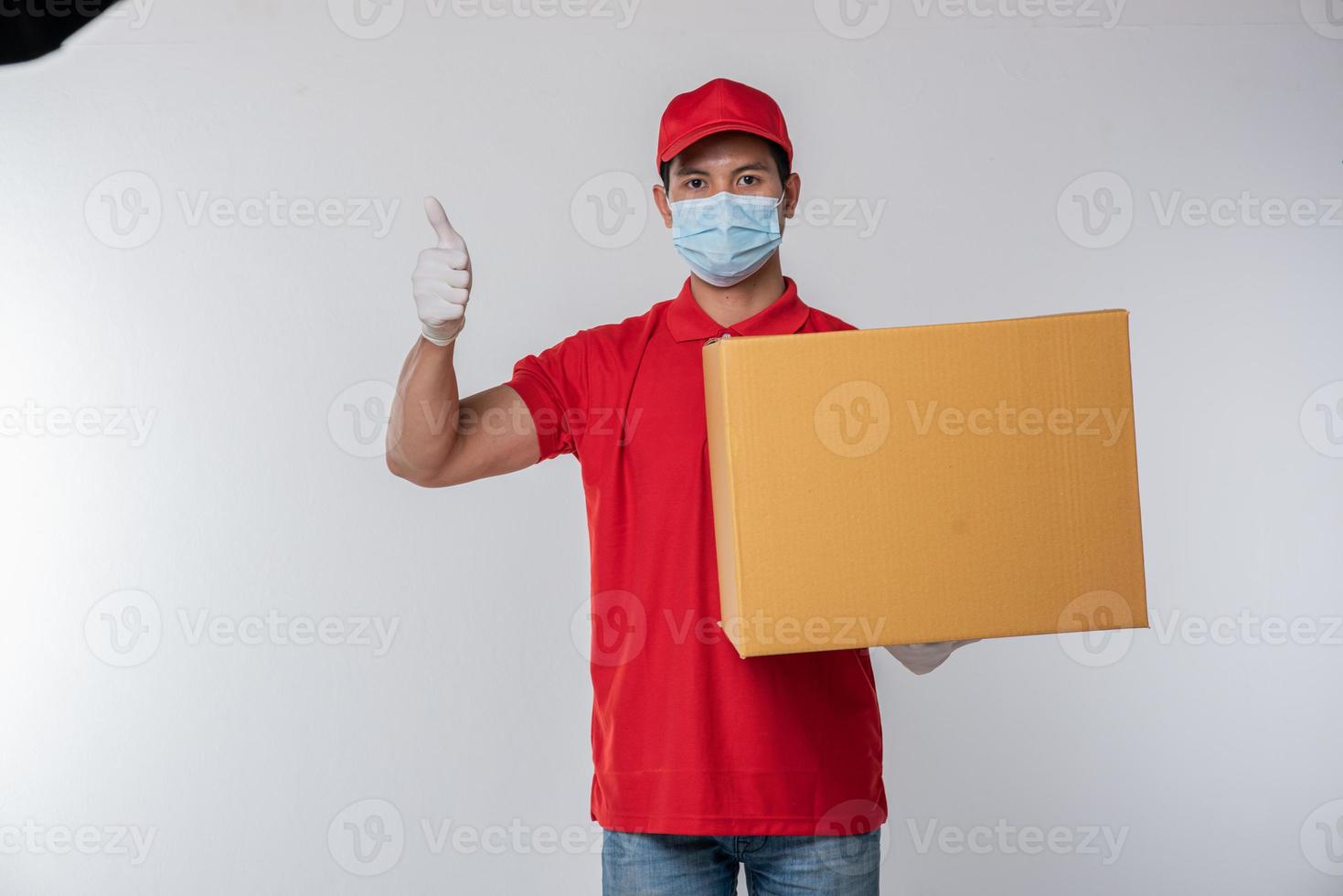 bild av en medveten ung leverans man i röd keps tom t-shirt enhetlig ansikte mask handskar stående med tömma brun kartong låda isolerat på ljus grå bakgrund studio foto