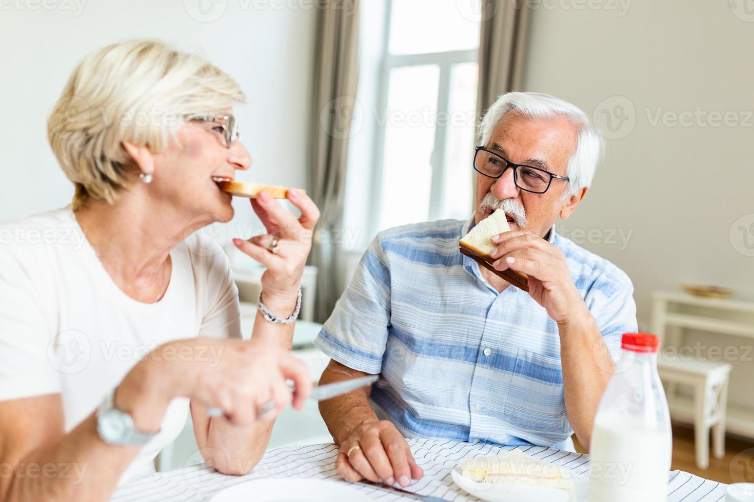 senior par äter frukost på Hem. innehav bit av bröd. äldre par njut av i deras tid tillsammans. känsla Lycklig. senior man äter smörgås foto