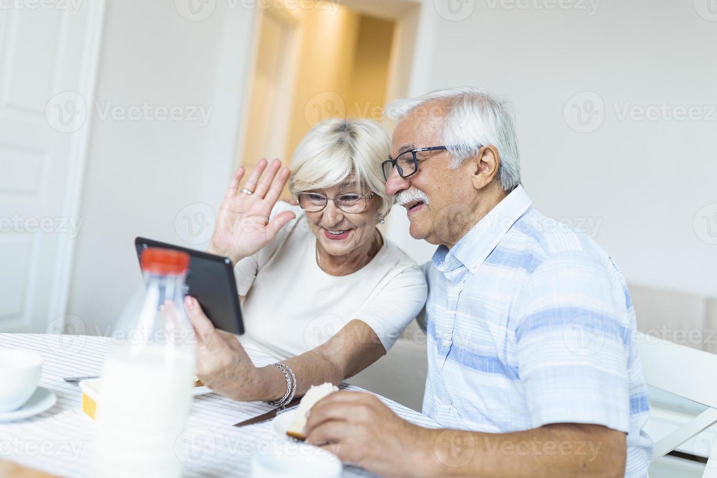 senior man och hans fru är använder sig av en digital läsplatta och kommunicerar med deras ungar. Lycklig senior par är har frukost och har ett uppkopplad konversation med deras familj och sändning hälsningar. foto