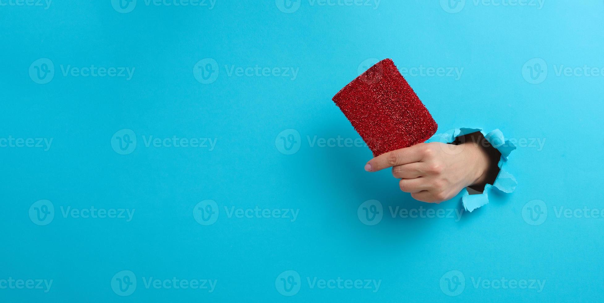 kvinna hand innehar en röd kök svamp. del av de kropp fastnar ut av en trasig hål i en blå papper bakgrund foto