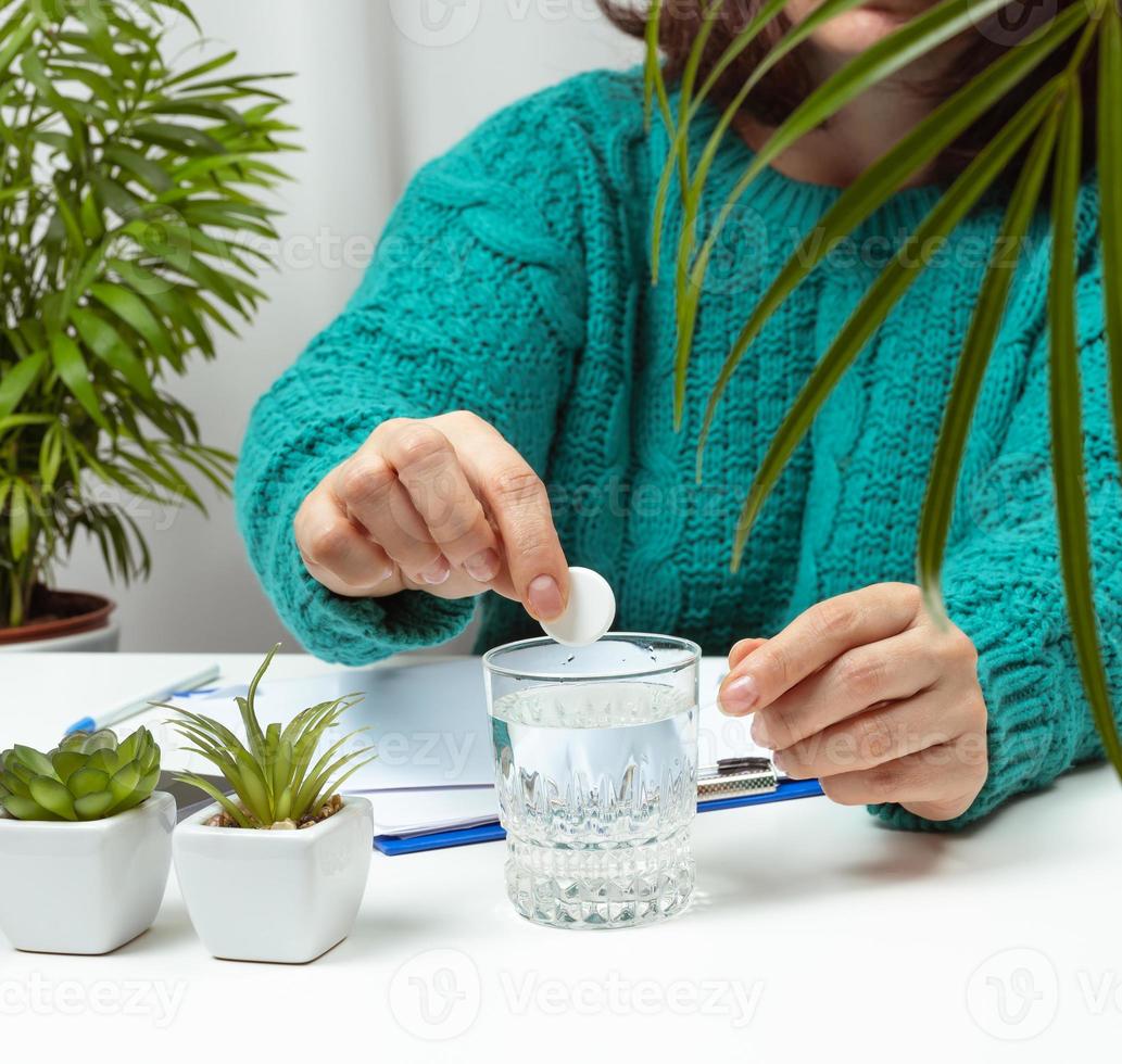 kvinna sitter på de tabell och kastar en runda aspirin in i de sakan med vatten, huvudvärk foto