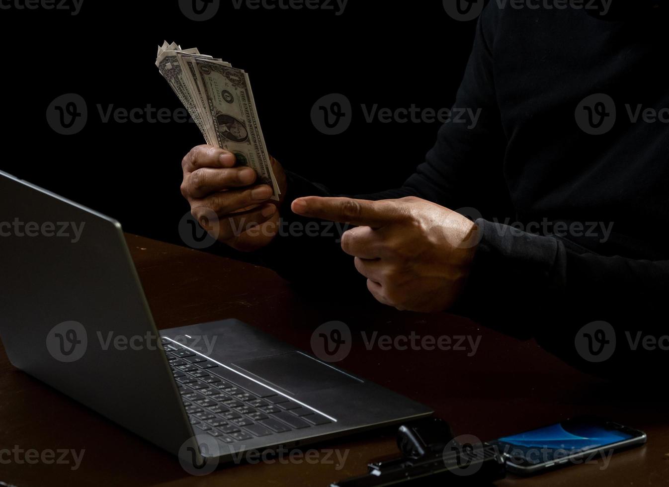 dator och händer av en man bär en svart skjorta, Sammanträde på en stol och en tabell, är en tjuv, innehav pengar, räkning de belopp erhållna från kapning eller råna, i en kolsvart rum. foto