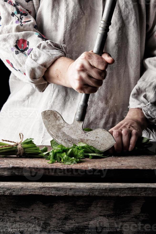 kvinna i en grå Linné klänning är skärande grön löv av färsk ängssyra på en trä- skärande styrelse med ett gammal metall kniv foto