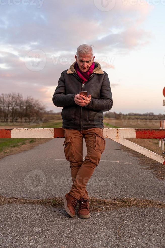 mitten åldrig man bär vinter- kläder använder sig av smartphone i en Land väg foto
