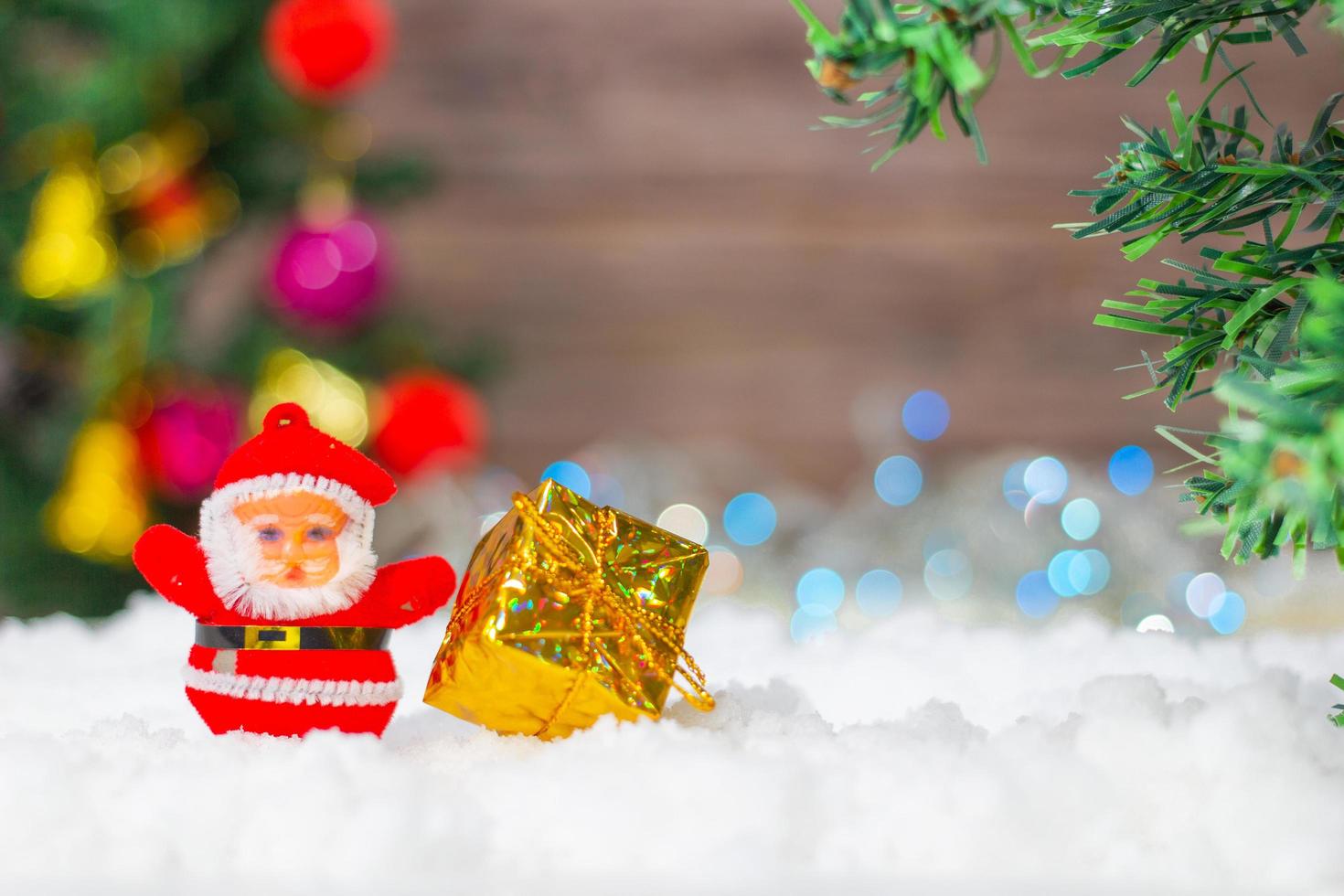 santa claus docka med en gåva låda placerad på de snö och de jul träd bakgrund med bokeh och trä. foto