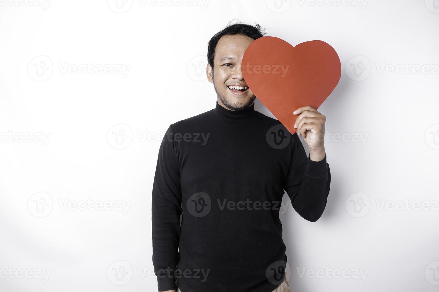 en porträtt av en Lycklig asiatisk man bär en svart skjorta, innehav en röd hjärtformade papper isolerat förbi vit bakgrund foto