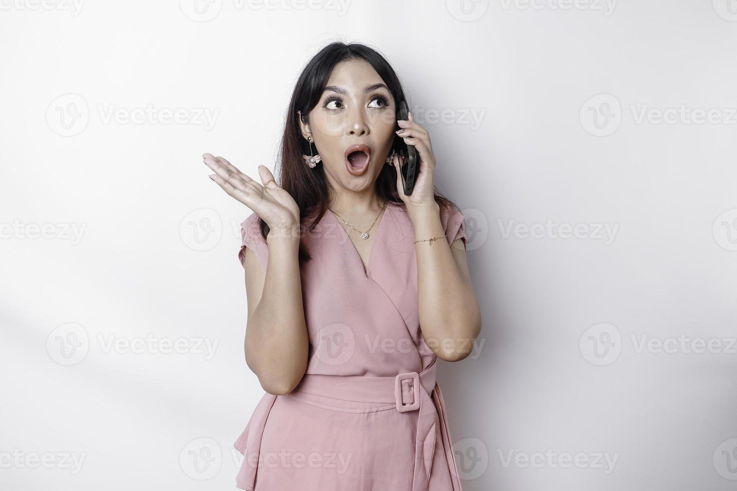 chockade asiatisk kvinna klädd i rosa och innehav henne telefon, isolerat förbi vit bakgrund foto