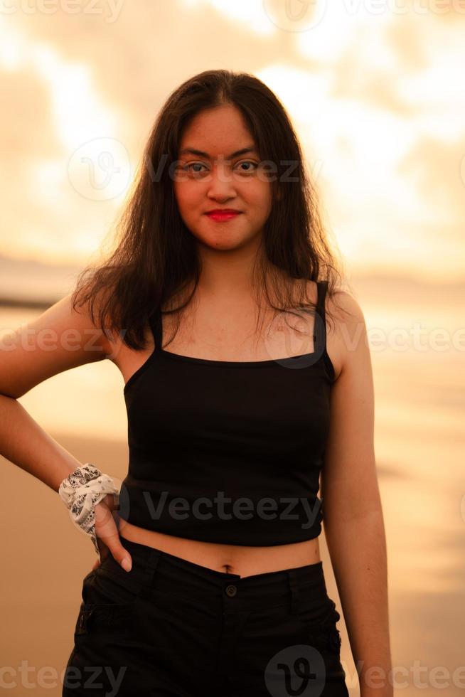 ett asiatisk Tonårs flicka i en svart skjorta med en leende uttryck står i främre av de sand eller med de vågor kraschar medan njuter de se foto