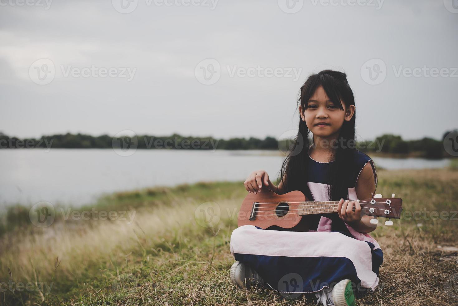 söt liten asiatisk tjej som sitter på gräset och spelar ukulele i parken foto