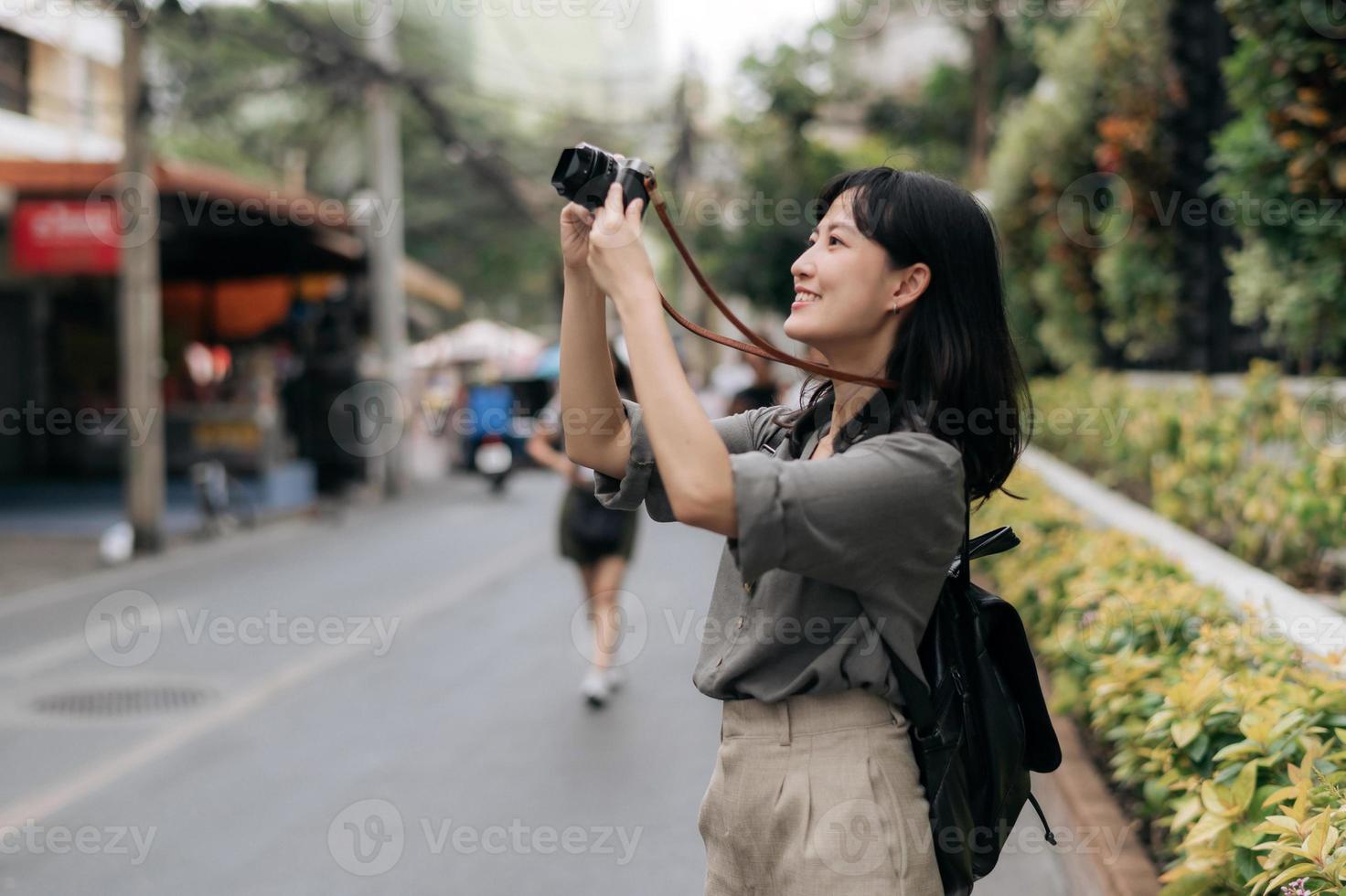 ung asiatisk kvinna ryggsäck resande använder sig av digital kompakt kamera, njuter gata kulturell lokal- plats och leende. resande kontroll ut sida gator. foto