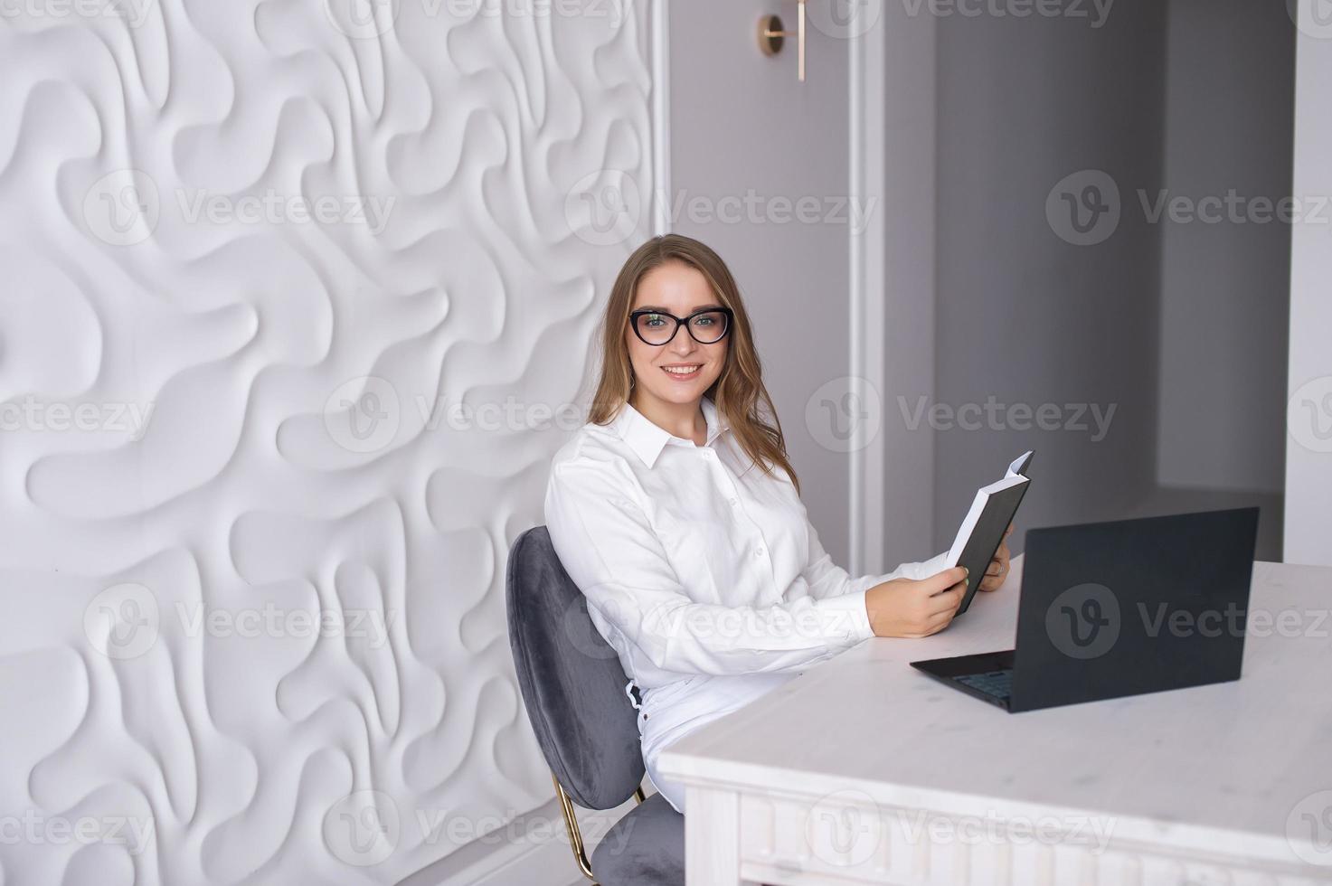 en ung lärare med glasögon innehar en dagbok i henne händer och sitter på en tabell med en bärbar dator foto