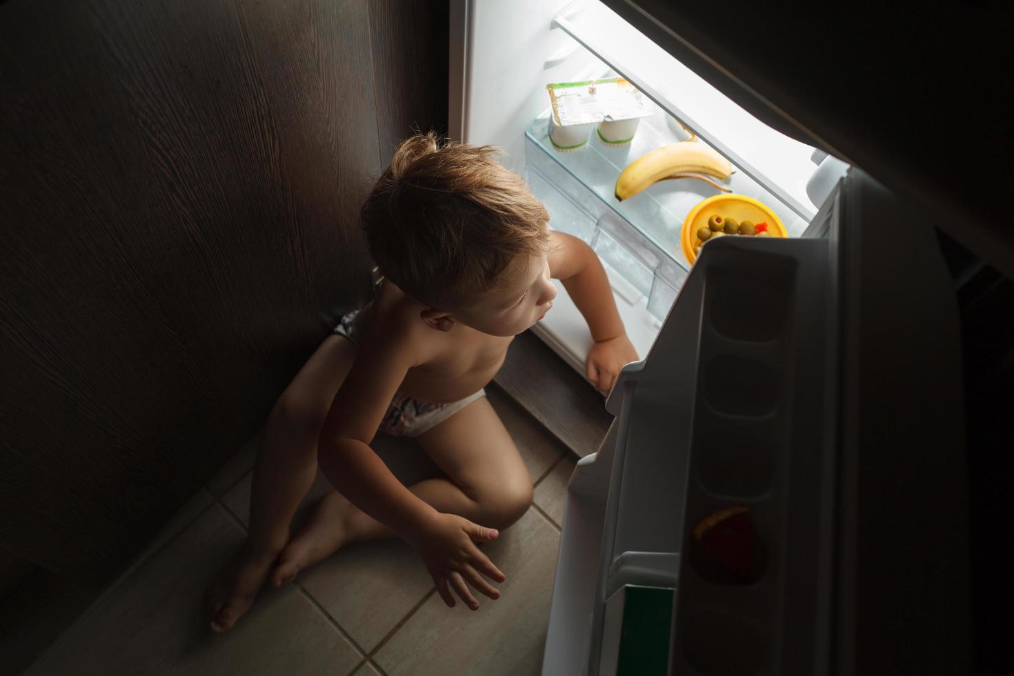 liten pojke som sitter nära ett öppet kylskåp på natten foto