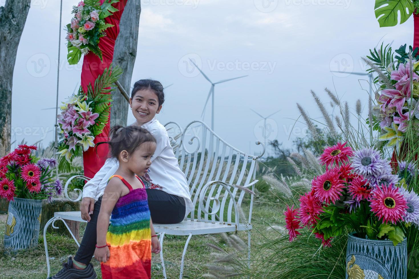 skön ung mor dotter sitter och koppla av på sommar äng bakgrund och väderkvarnar, väderkvarnar som alternativ energi källor, hållbar resurs begrepp, gemenskap människor med väderkvarnar foto