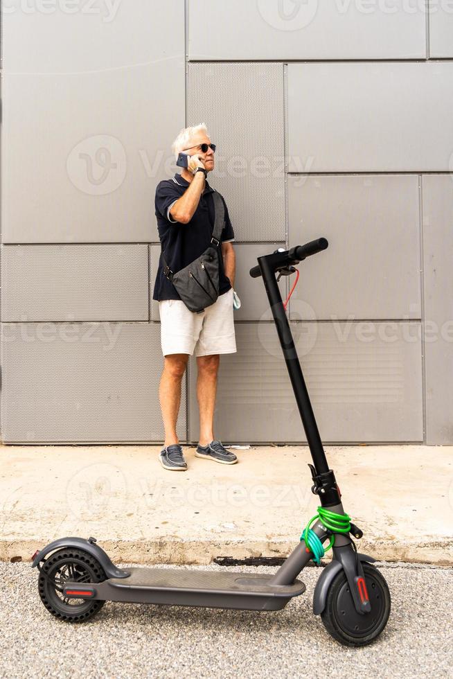 pendlare hipster man med elektrisk skoter är använder sig av smartphone i de förorter foto