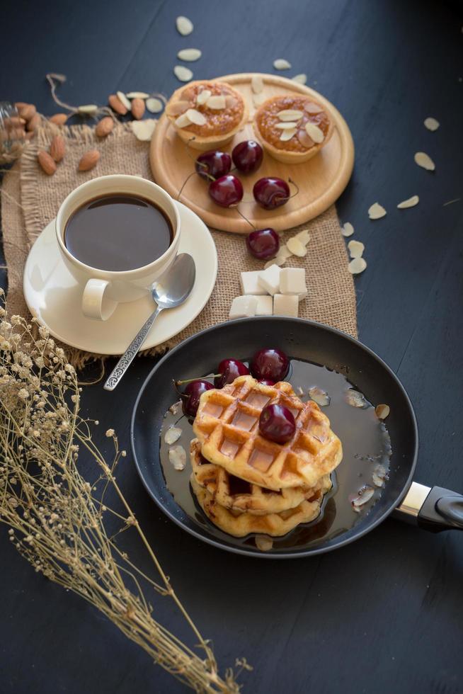 våfflor och körsbär med honung, krispiga mandeltårtor och en kopp kaffe på det svarta bordet foto