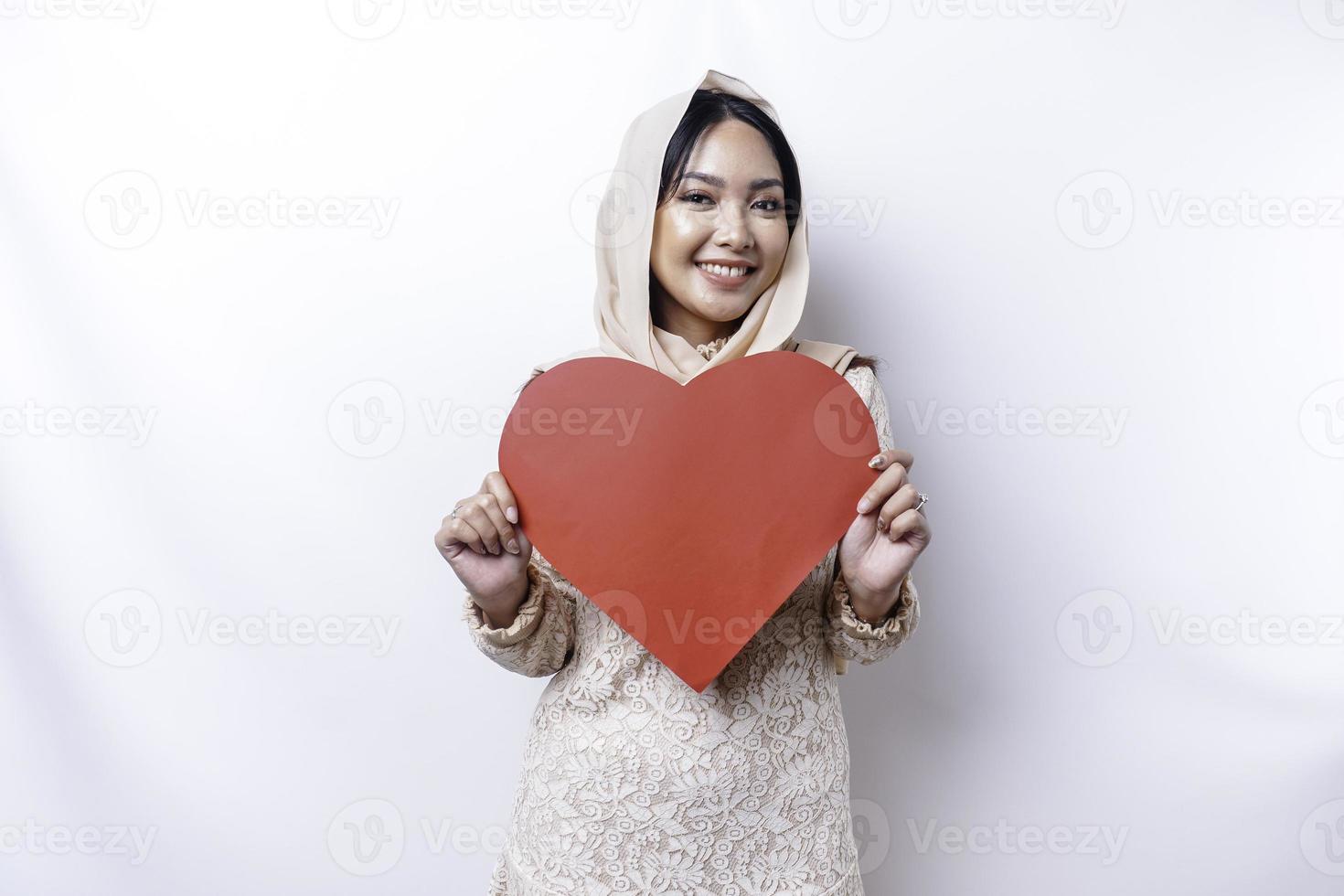 en Lycklig ung asiatisk muslim kvinna bär en hijab känner romantisk former hjärta gest uttrycker anbud känslor och innehav en röd hjärtformade papper foto