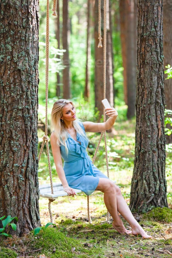 skön blond kvinna i blå klänning använder sig av smartphone till tar selfie foto