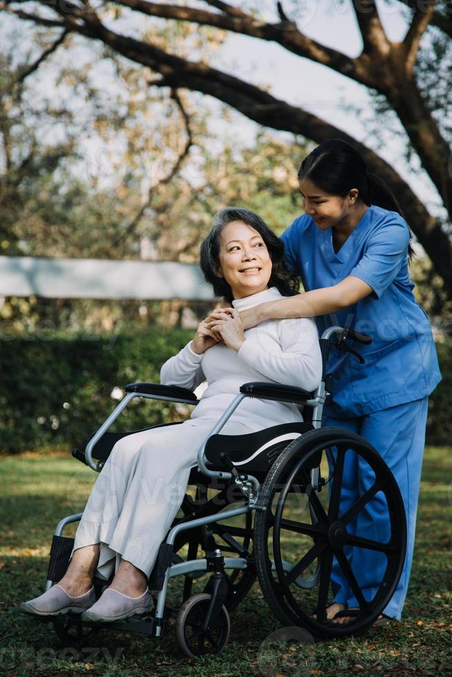 asiatisk ung vårdgivare sjuksköterska Stöd senior äldre manlig gående utomhus. specialist flicka läkare hjälp och ta vård av äldre mogna man patient håller på med fysisk terapi i offentlig parkera på amning Hem. foto