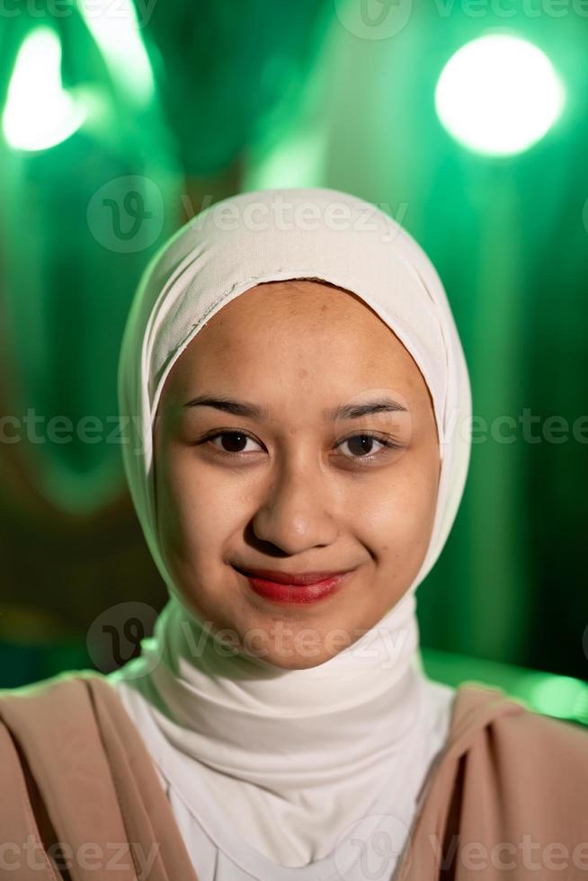 en muslim kvinna med en vit slöja och vit kläder leende mycket Söt utan smink på henne ansikte i en grön rum foto