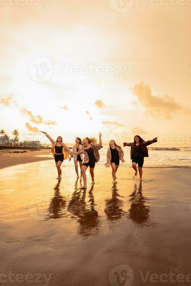 en grupp av asiatisk tonåringar i shirts löpning med deras vänner med mycket glad uttryck på de strand foto