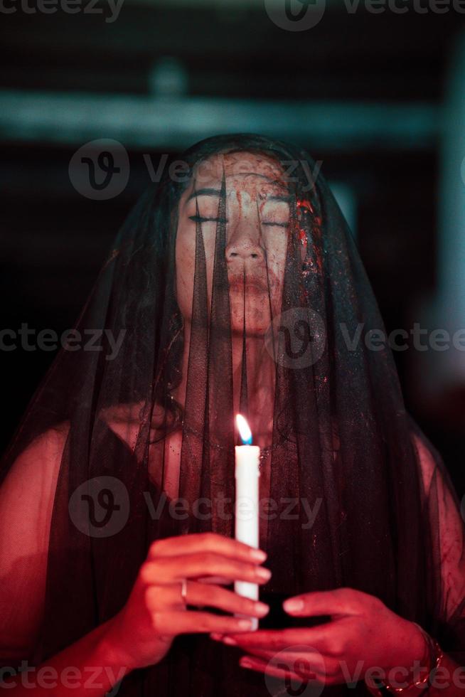 ett asiatisk kvinna med stängd ögon och en transparent slöja färgade med blod innehar en ljus i henne hand under satanisk dyrkan foto