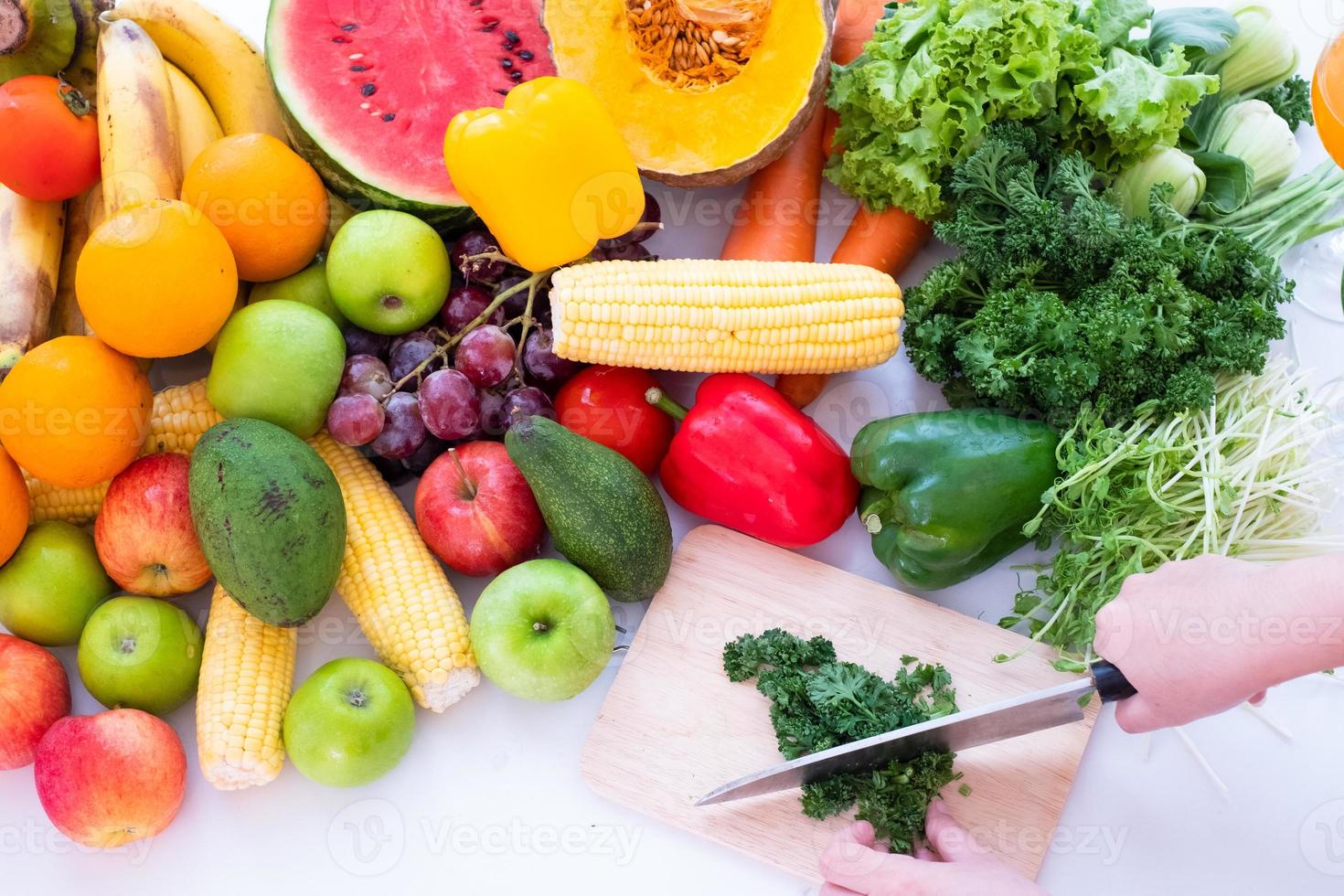 en personens händer är synlig innehav en kniv och hackning grönsaker på en skärande styrelse med frukt de bakgrund är en vit ridå foto