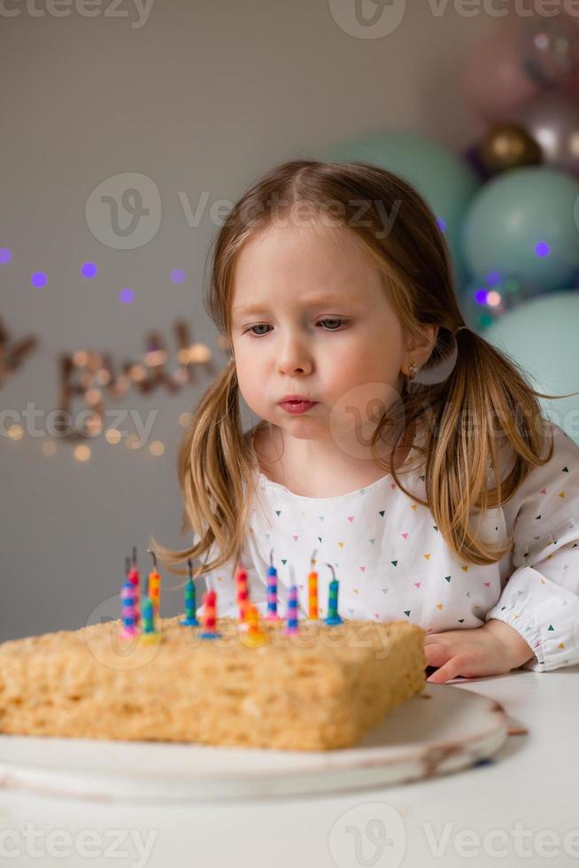 söt liten flicka slag ut ljus på en födelsedag kaka på Hem mot en bakgrund av ballonger. barnets födelsedag foto