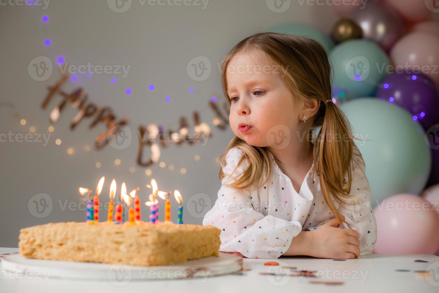 söt liten flicka slag ut ljus på en födelsedag kaka på Hem mot en bakgrund av ballonger. barnets födelsedag foto