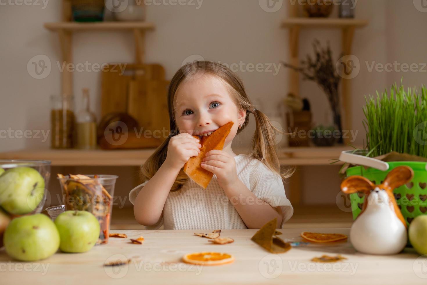 söt liten flicka äter naturlig pastill på Hem i en trä- kök. mat för barn från naturlig Produkter foto