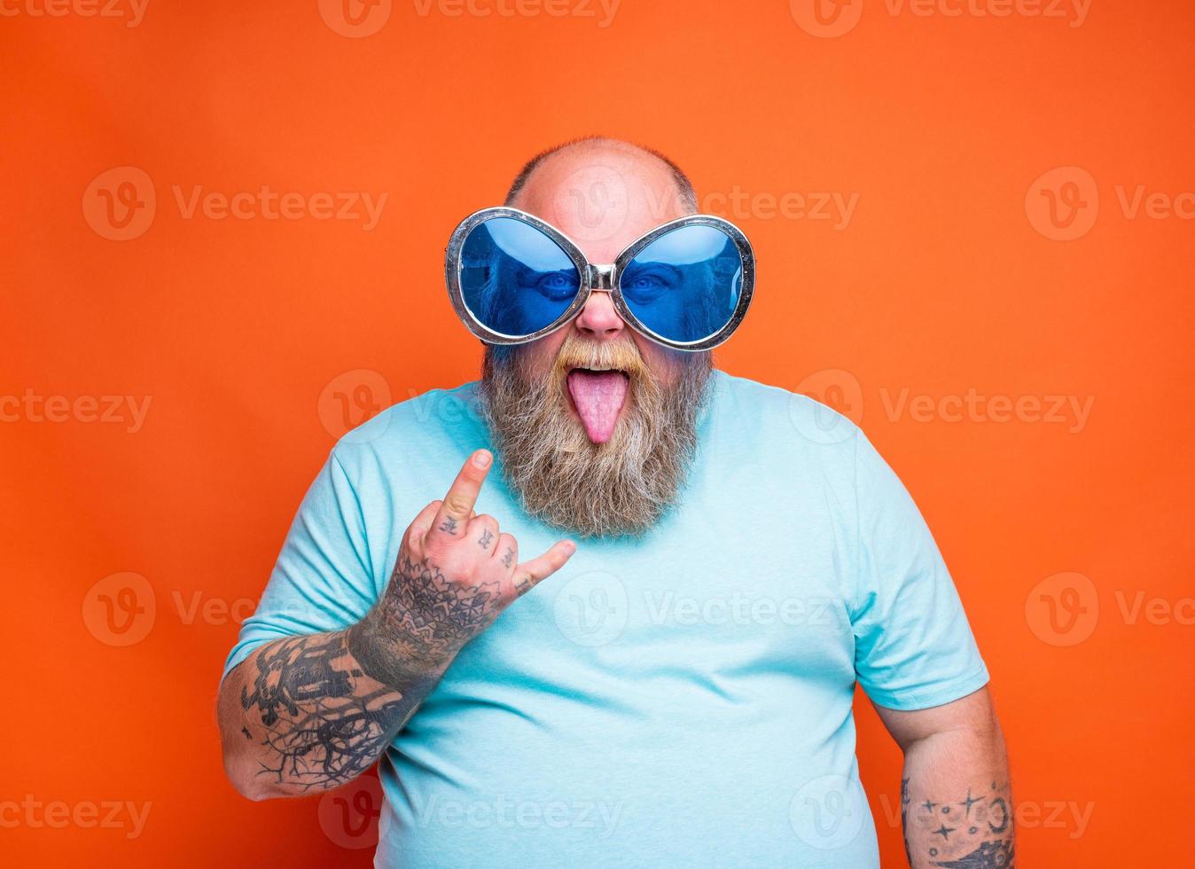 fett man med skägg, tatueringar och solglasögon gör de gest av de horn foto