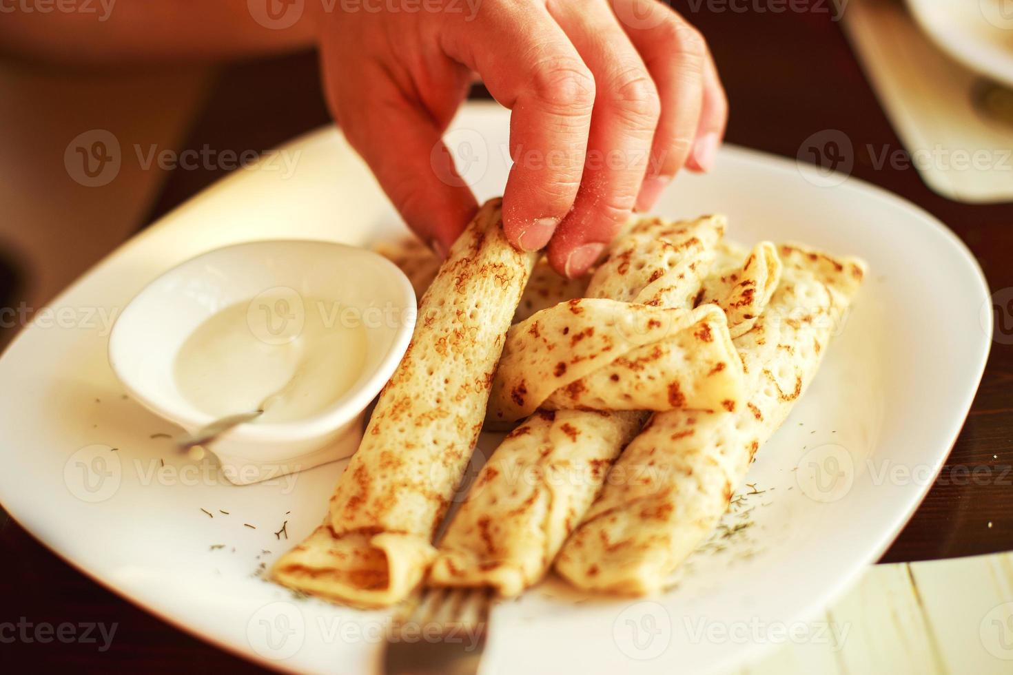 pannkakor rullad i en sugrör på en tallrik med sur grädde. man äter pannkaka foto