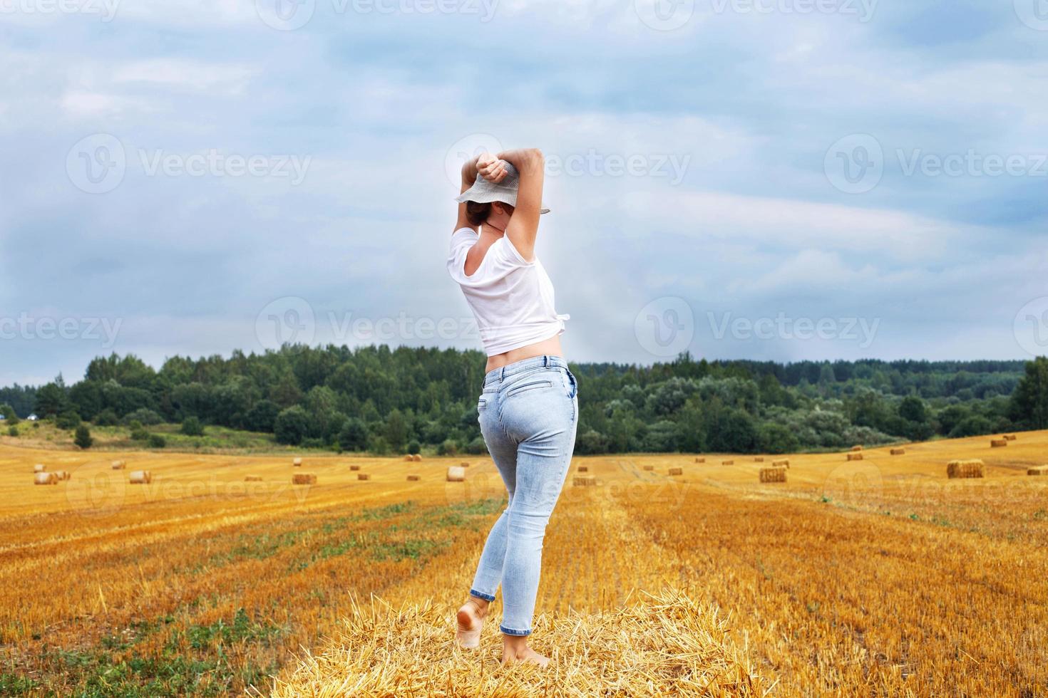 flicka i sugrör hatt står på en höstack på en bal i de jordbruks fält efter skörd foto