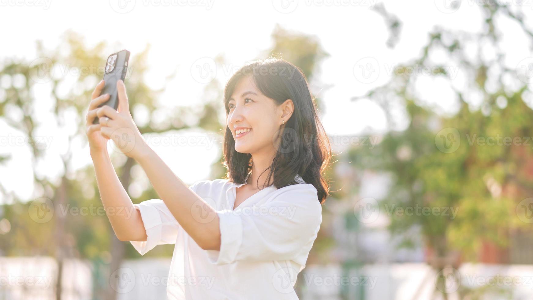 porträtt skön ung asiatisk kvinna med smart mobil telefon runt om utomhus- natur se i en solig sommar dag foto