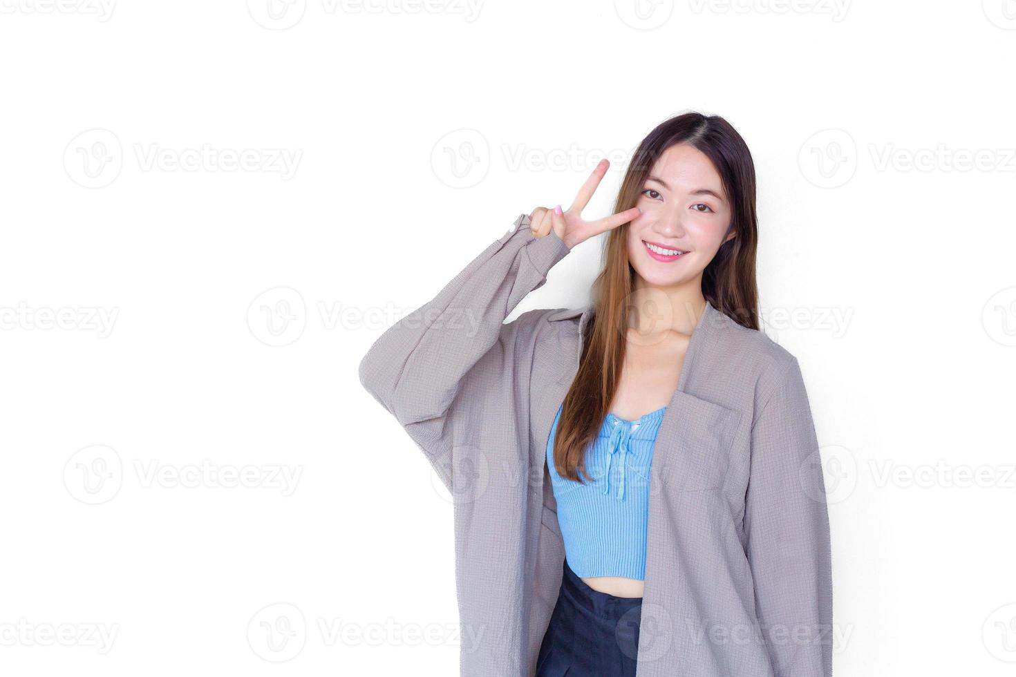 skön ung asiatisk kvinna lång brun hår i blå skjorta är är stående självsäkert framgångsrik verkande hand visar två tummen upp som tecken av seger som presenter något isolerat på vit bakgrund. foto