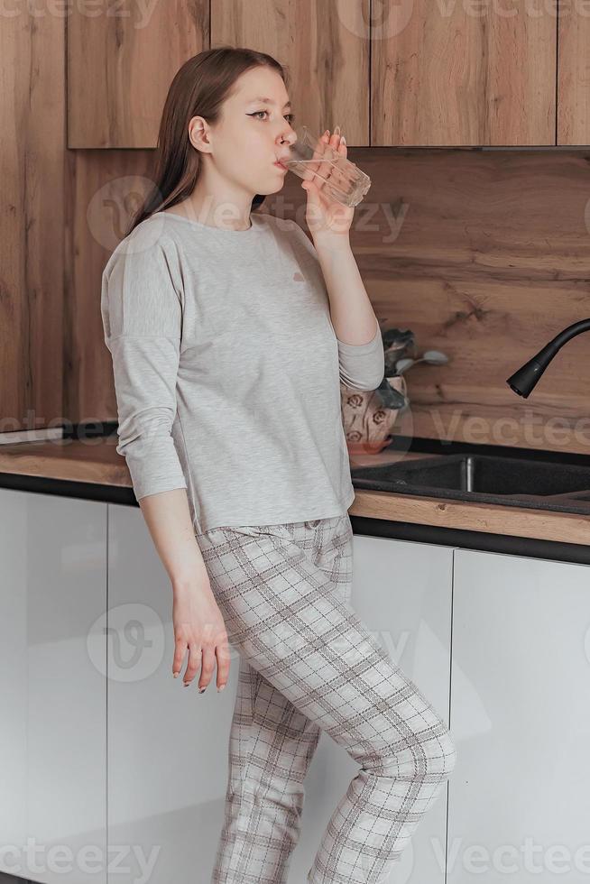 en ung skön flicka i pyjamas drycker rena vatten i de kök i de morgon. de begrepp av en friska livsstil, de fördelar av morgon- vatten på ett tömma mage. foto