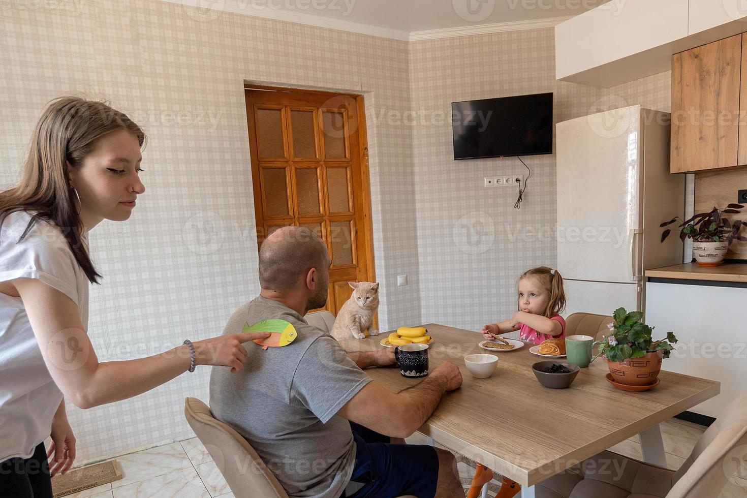 de familj under frukost i de kök på de först av april skämt och pinnar papper fisk på varje andras ryggar. rolig familj skämt, traditioner, april dåre dag firande foto