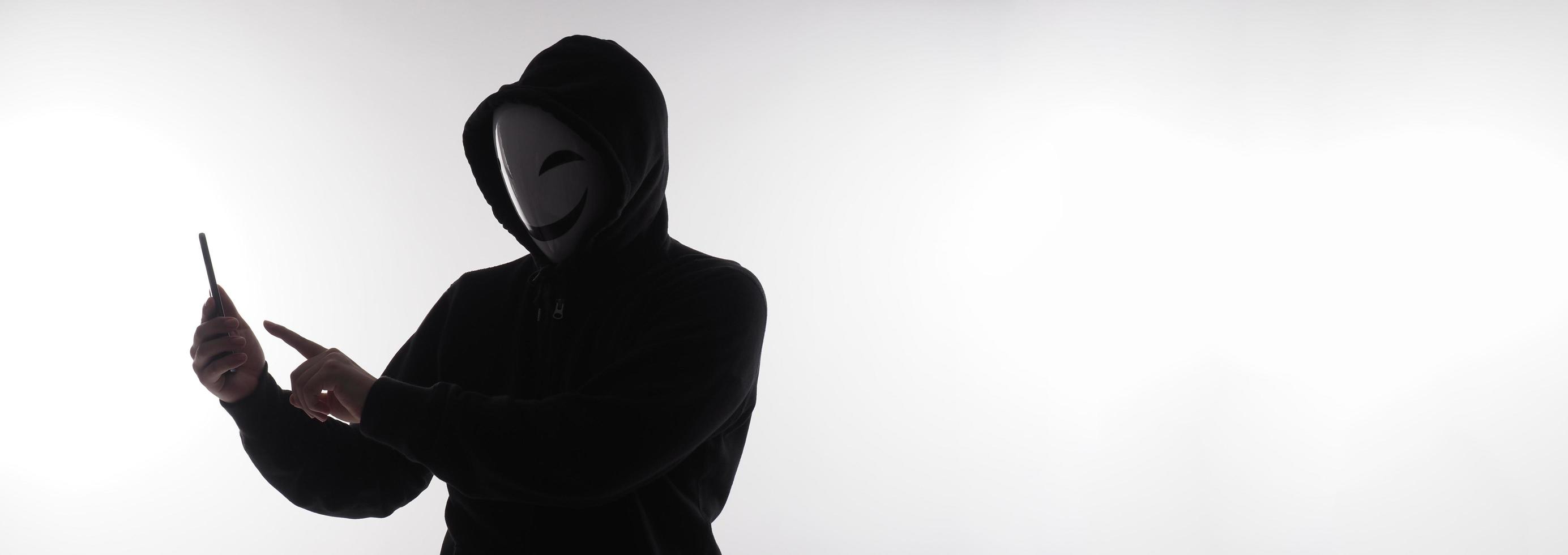 hacker anonym och ansikte mask med smartphone i hand. man i svart huva skjorta innehav och använder sig av mobil telefon på vit bakgrund. representera cyber brottslighet data dataintrång eller stjäla personlig data begrepp foto