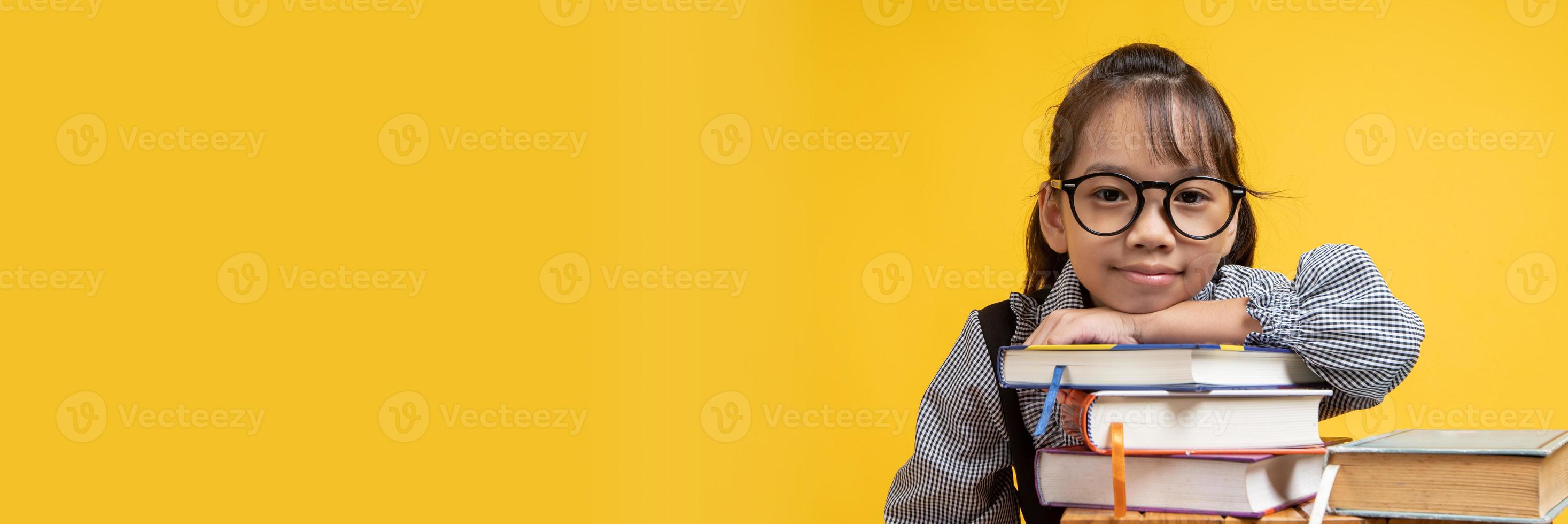 thailändsk asiatisk tjej som bär glasögon lutar på buntböcker och ser kamera med gul bakgrund foto