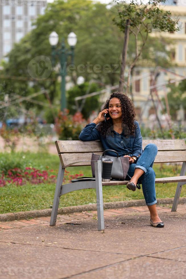 ung afrikansk amerikan affärskvinna talande på de telefon Sammanträde på de bänk i de stad parkera foto