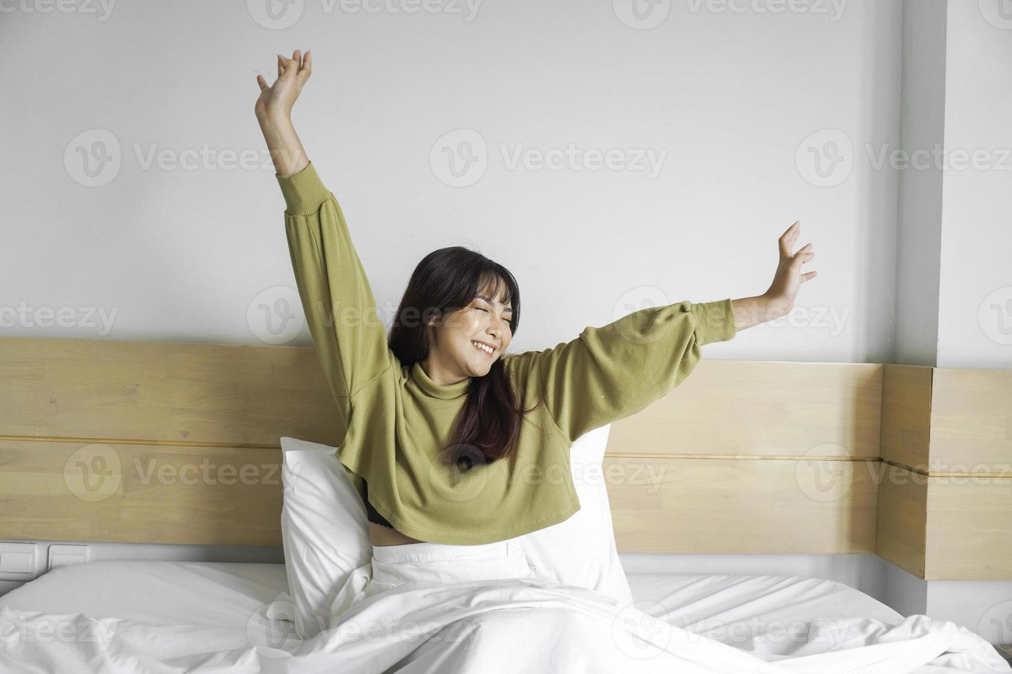 porträtt av skönhet Lycklig asiatisk kvinna vakna, vakna upp på de säng. ung skön asiatisk flicka stretching henne vapen i färsk tidigt morgon, koppla av fredlig morgon- rutin- gäspa begrepp baner foto