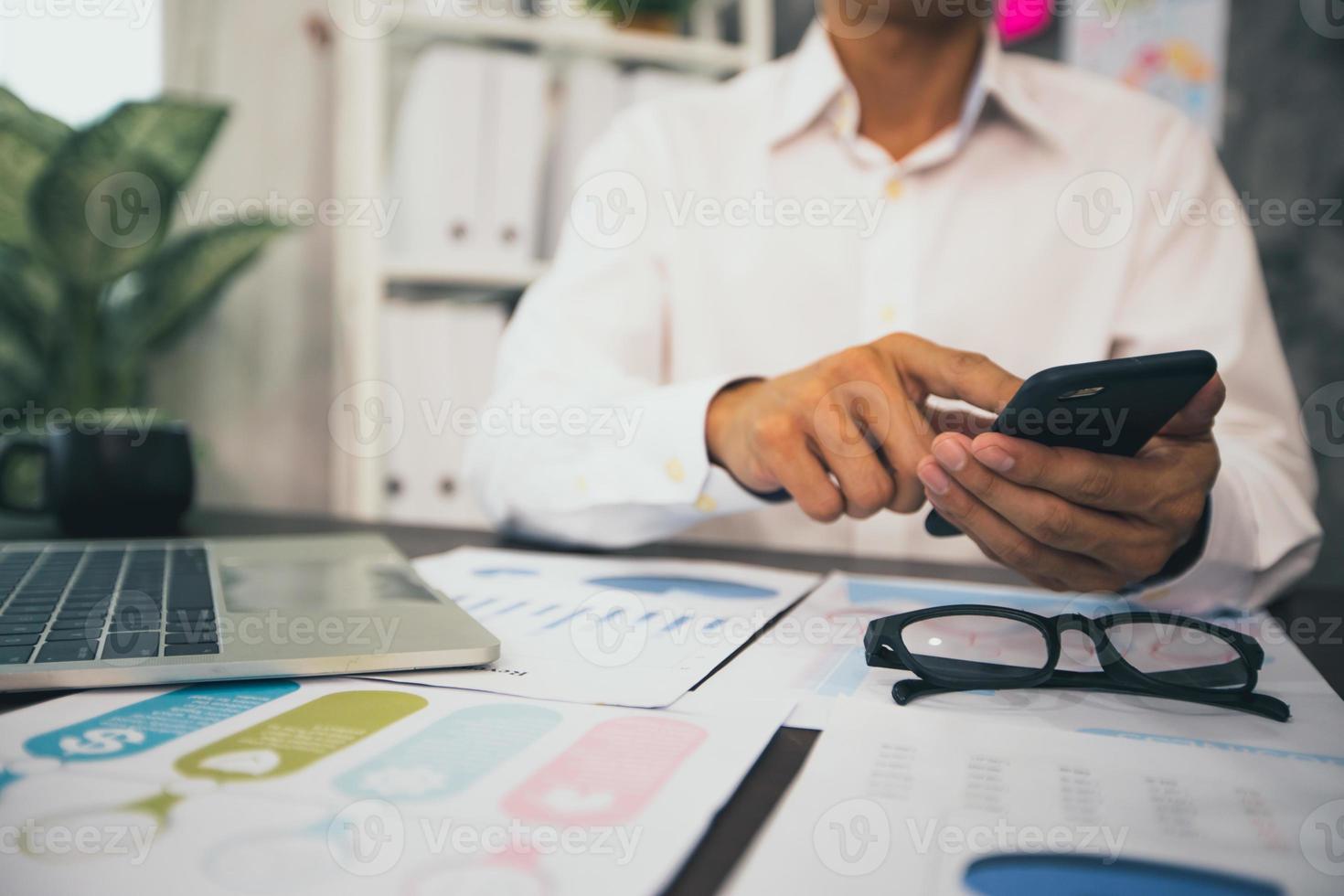 affärsman som arbetar på mobiltelefon bredvid bärbar dator med papper av diagram och grafer foto