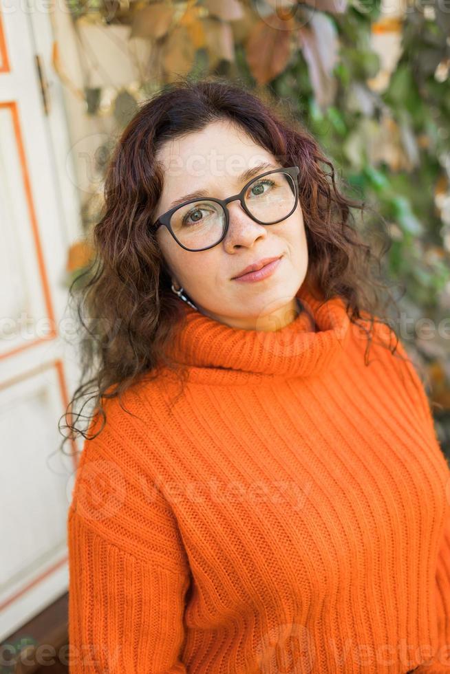 höst porträtt av ett attraktiv ung kvinna i eleganta glasögon i en stickat modern orange Tröja på bakgrund av löv. flicka promenader i stad foto