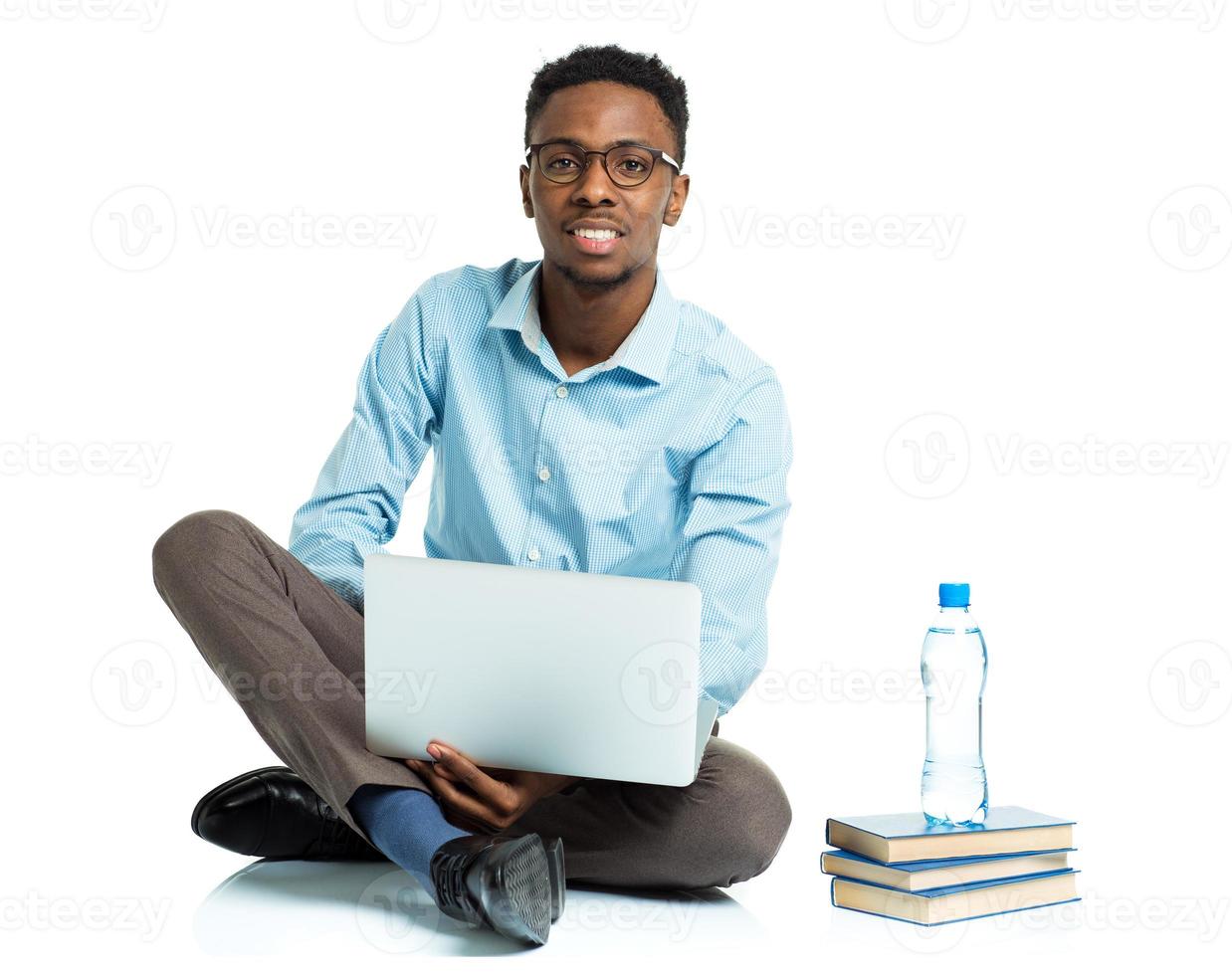 Lycklig afrikansk amerikan högskola studerande med bärbar dator, böcker och flaska av vatten Sammanträde på vit foto