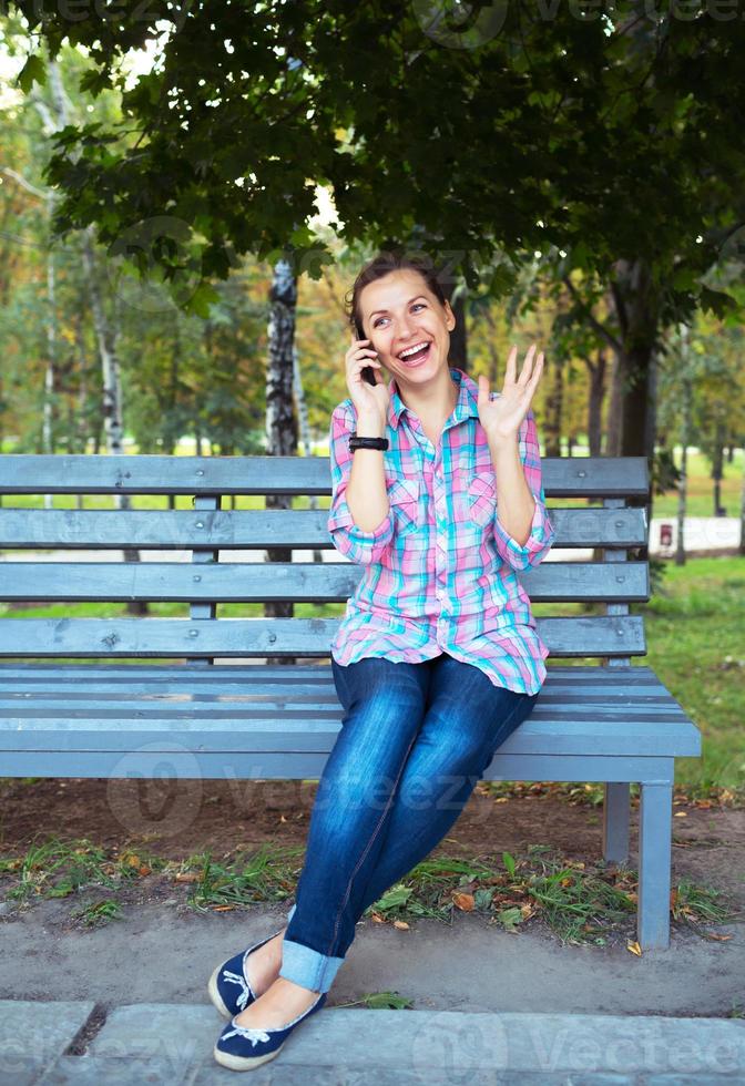 en porträtt av en leende kvinna i en parkera på en bänk talande på de telefon foto