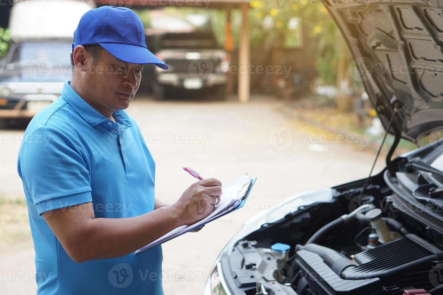 asiatisk man mekaniker bär blå keps och blå skjorta, innehar papper, kontroll och analyserar bil motor under de huva. begrepp, utomhus- bil inspektion service. krav för olycka försäkring. foto